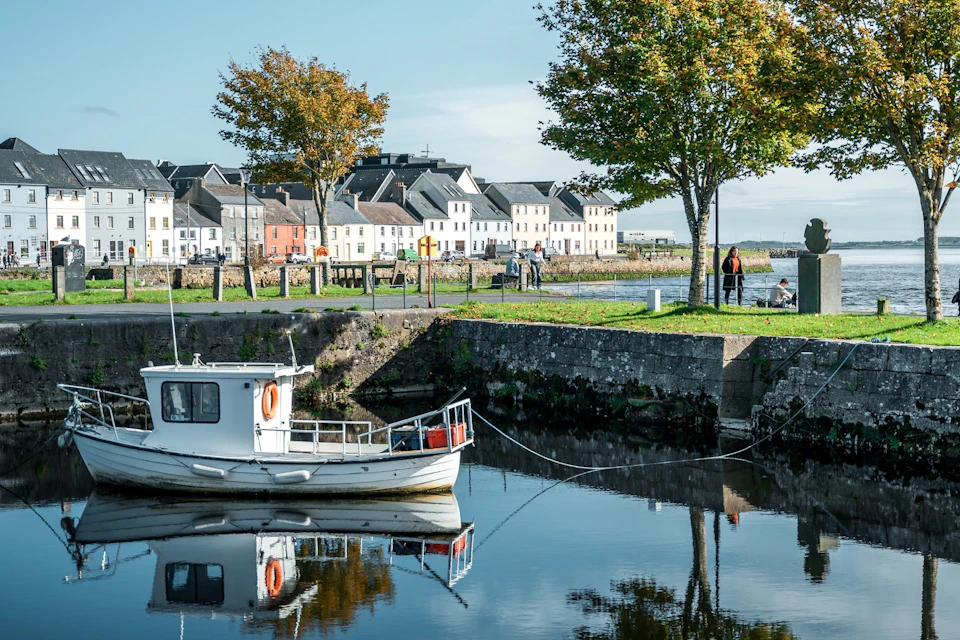 A small white boat docked on calm water near a stone wall, with trees, a green lawn, and colorful houses in the background.