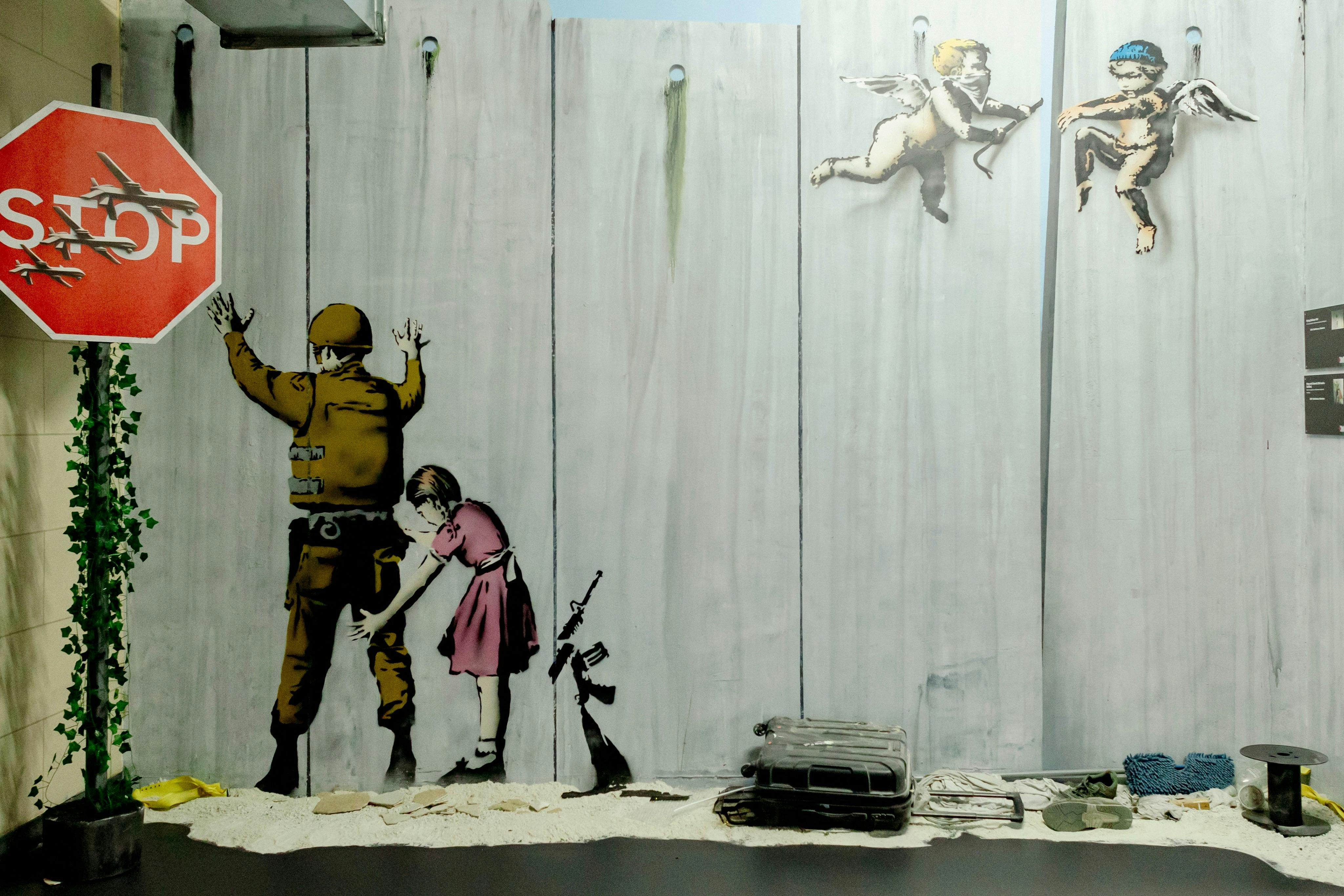 A mural of a young girl frisking a soldier on a wall. Two cherubs fly above, and a rifle leans against the wall.