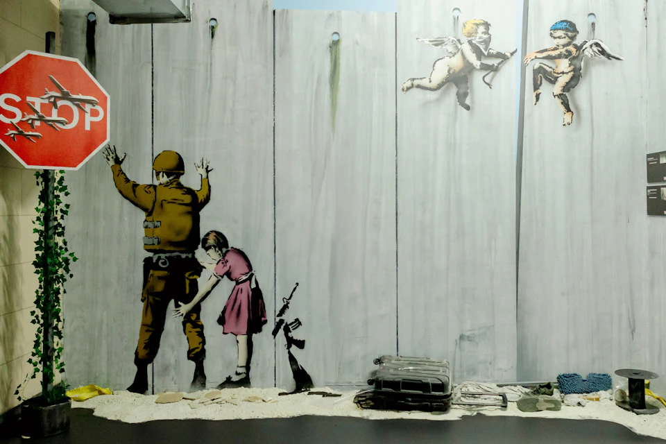 A mural of a young girl frisking a soldier on a wall. Two cherubs fly above, and a rifle leans against the wall.