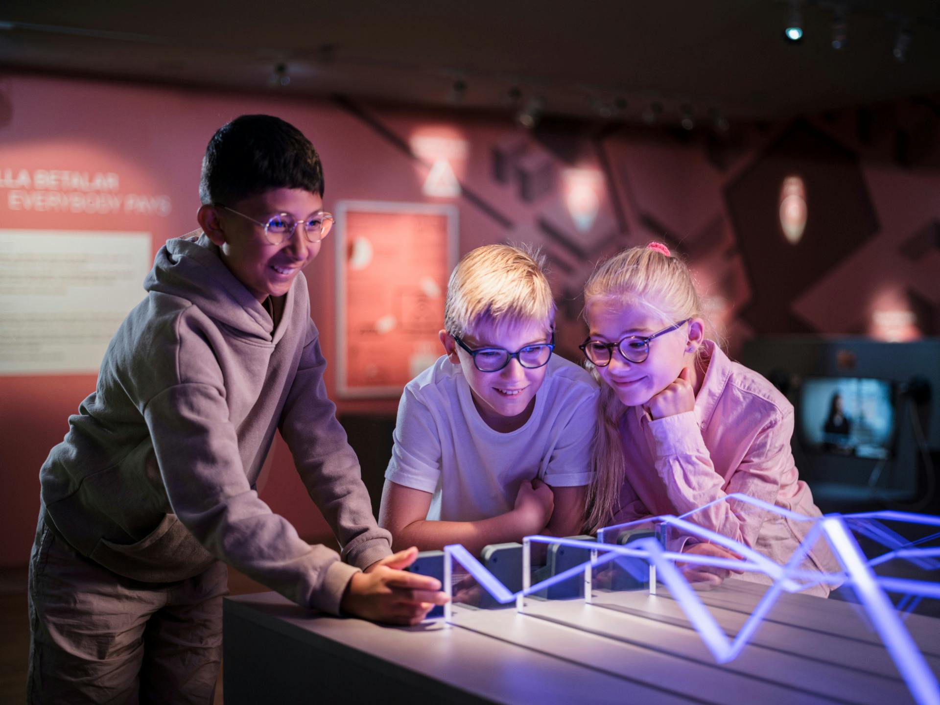 Drei Kinder interagieren mit einem beleuchteten Ausstellungsmodell in einer museumsähnlichen Umgebung, lächeln und sind beschäftigt.