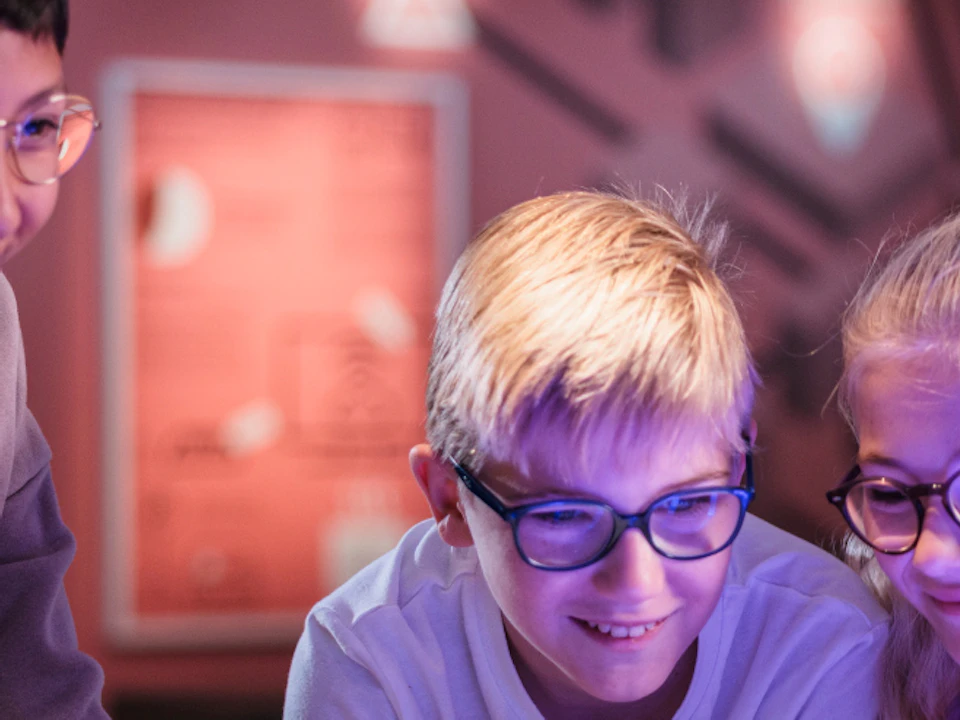 Three children interact with a lit-up model exhibit in a museum-like setting, smiling and engaged.