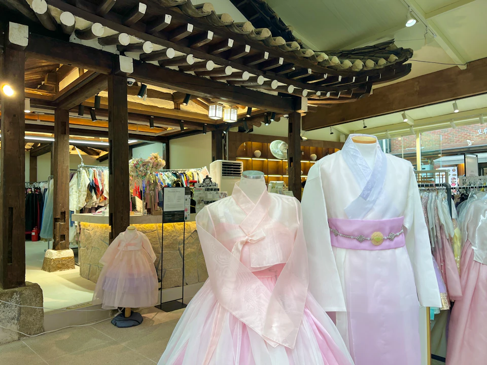 Traditional Korean hanbok dresses displayed on mannequins in a store with wooden decor and racks of colorful clothing.