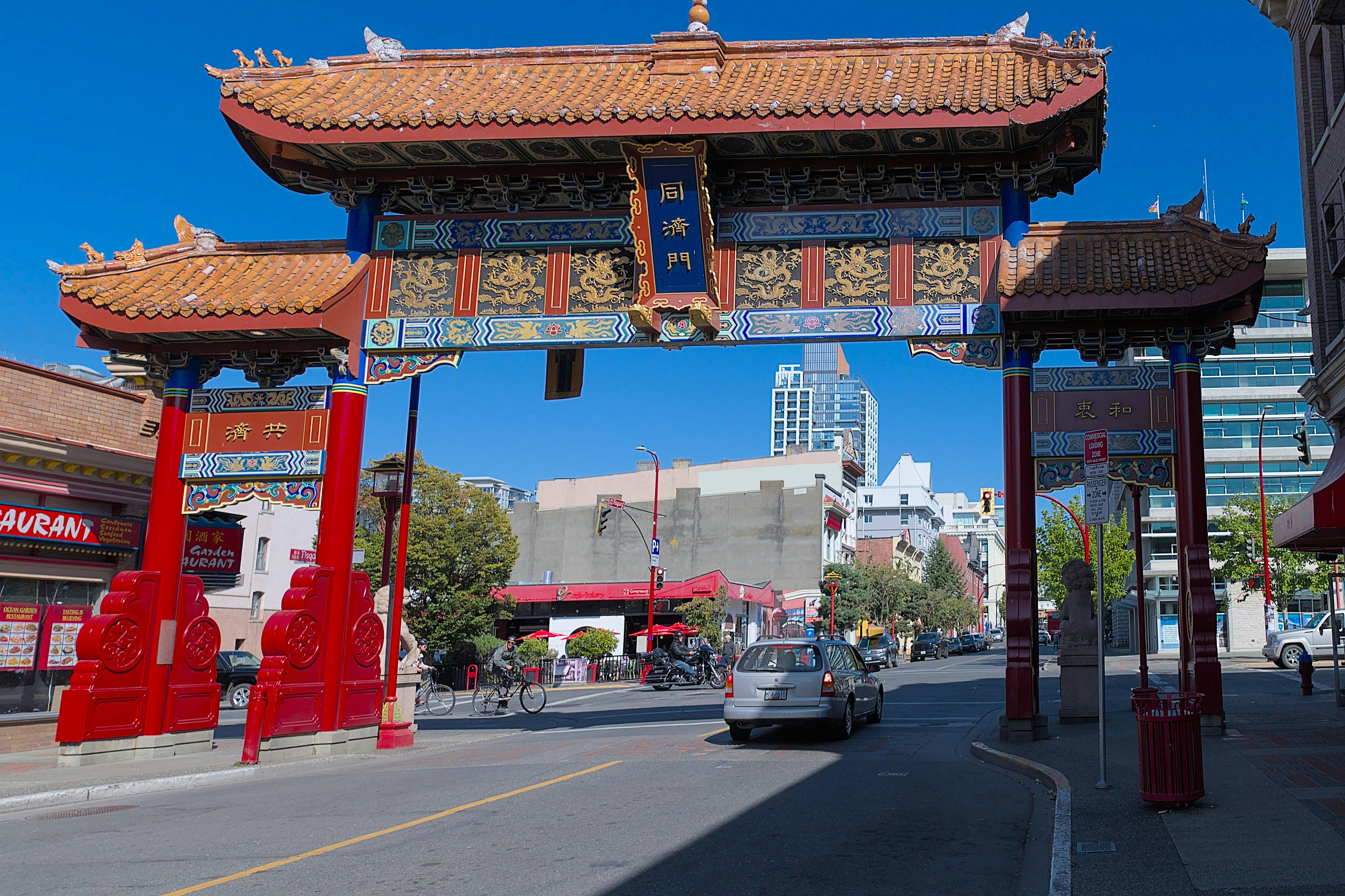 Μια παραδοσιακή κινεζική πύλη στέκεται πάνω από έναν δρόμο με αυτοκίνητα και κτίρια στο παρασκήνιο μια ηλιόλουστη μέρα.