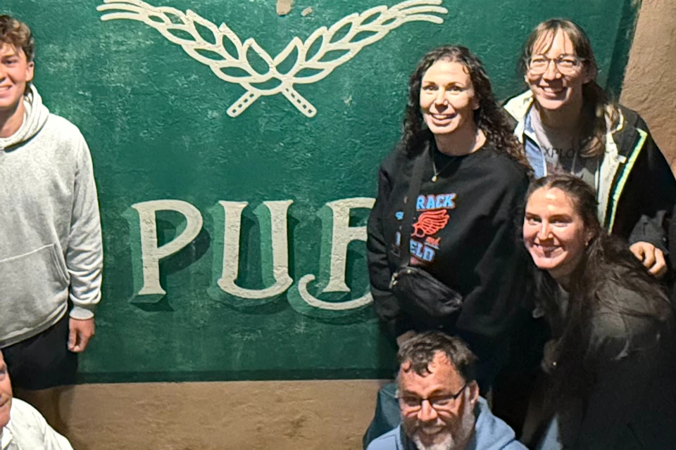 A group of people posing in front of a green sign that reads 'Harts Pub' with a decorative motif beneath the text.
