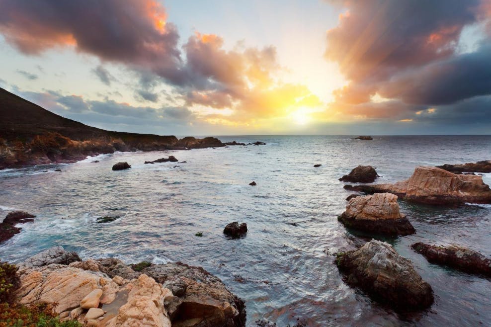 Skalnaté pobřeží při západu slunce s mraky, pronikajícími slunečními paprsky a vlnami narážejícími na velké skály v moři.