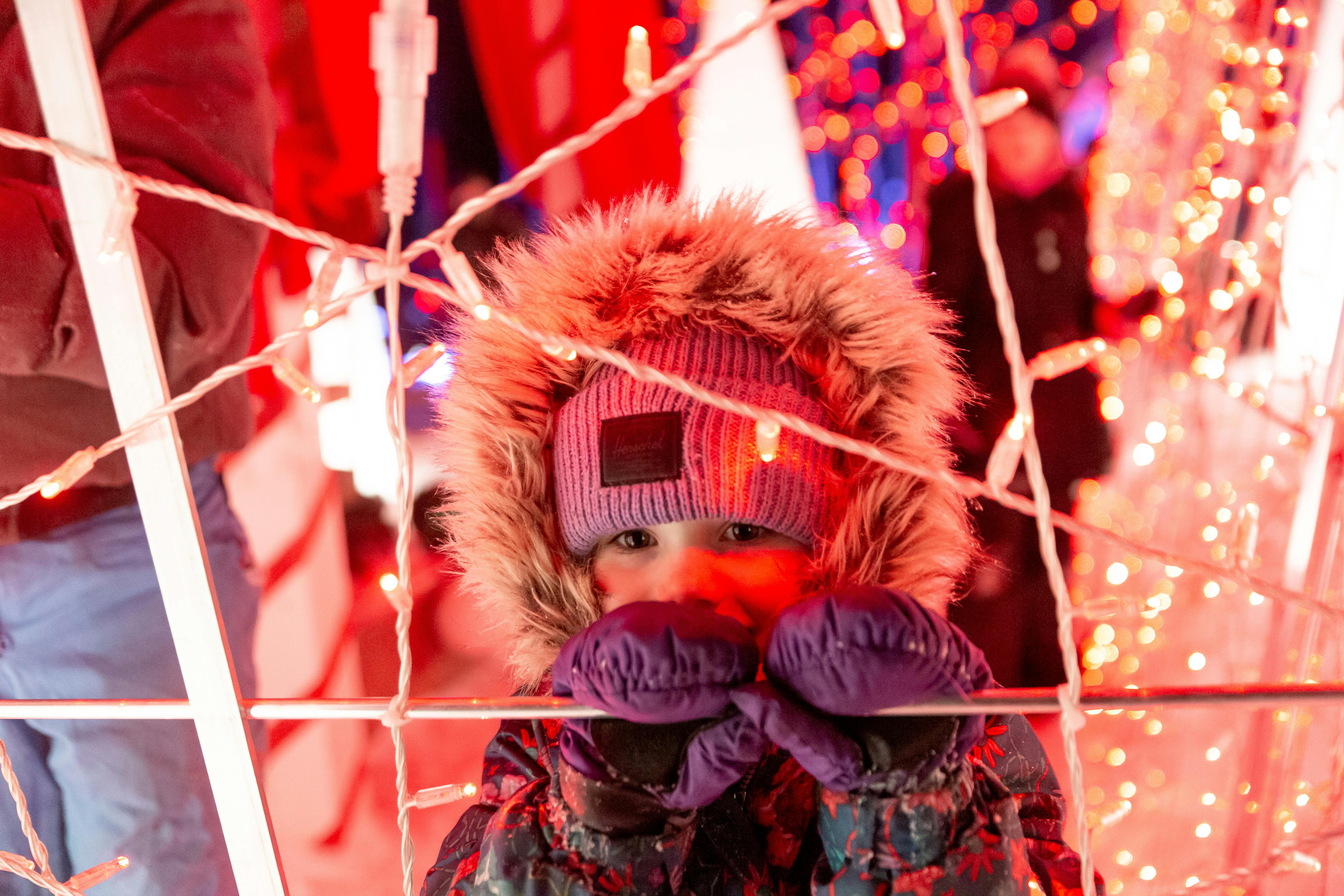 Een kind met een jas met capuchon en een paarse muts gluurt door de feestverlichting en houdt met gehandschoende handen een staaf vast.