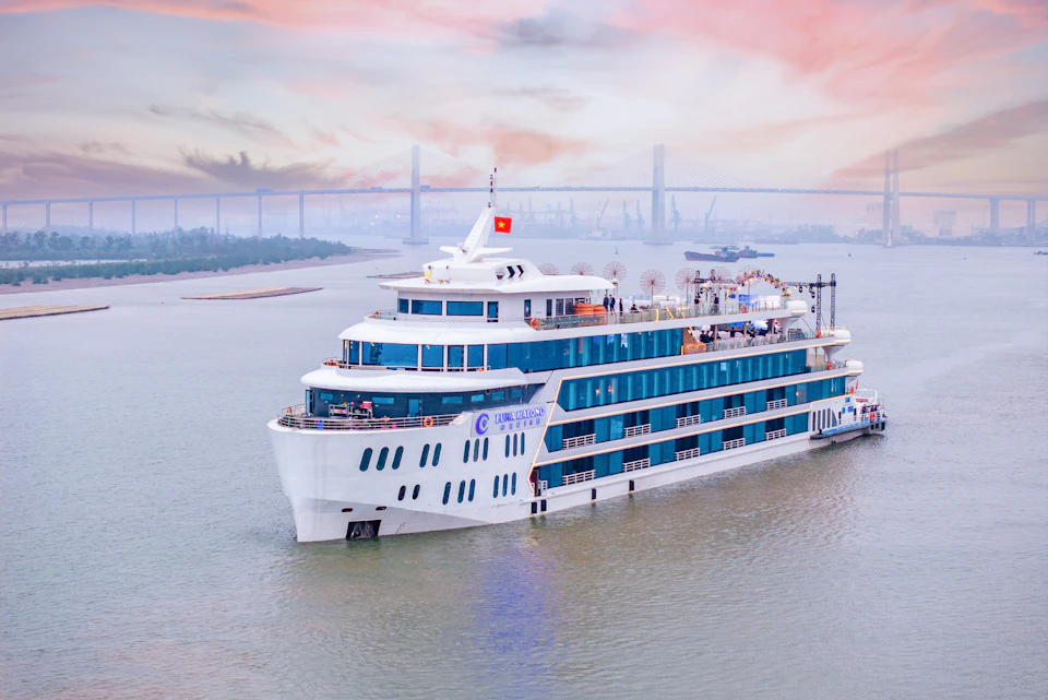 Large white cruise ship with blue windows and multiple decks sailing on a river with a bridge and sunset sky in the background.