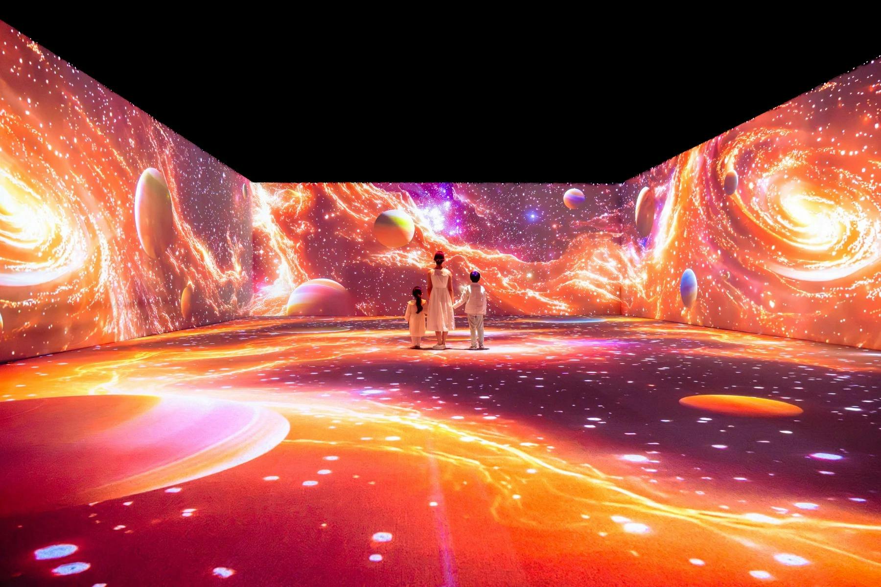三个人站在一个身临其境的房间里，房间里有生动的、以宇宙为主题的行星、恒星和旋转的星云投影。