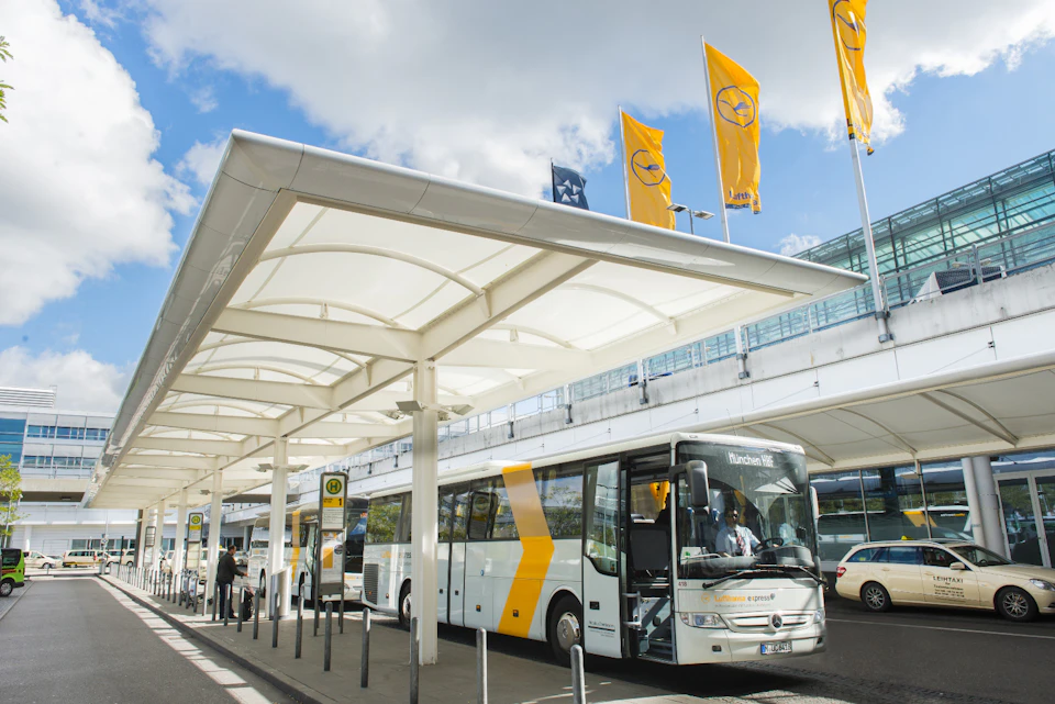 A white and yellow bus parked at an airport terminal under a covered area with yellow flags in the background.