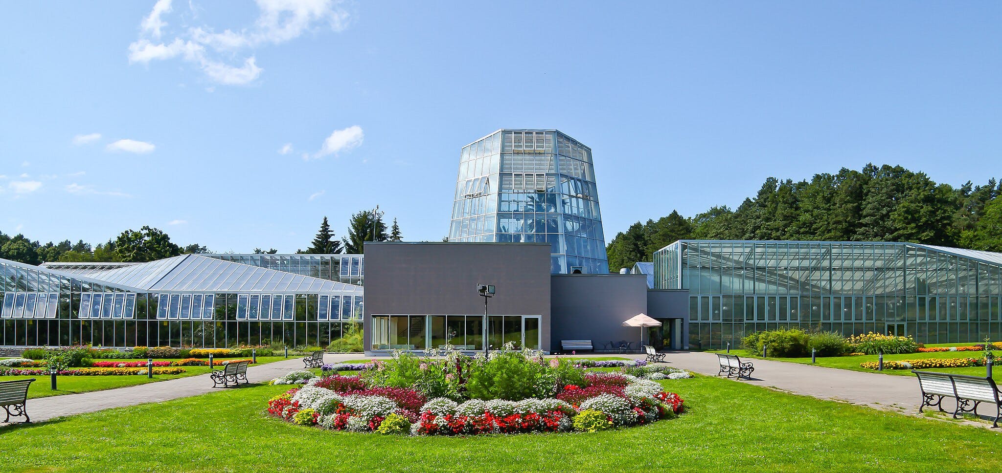A large modern greenhouse with a central glass tower, surrounded by landscaped gardens, benches, and paths under a clear sky.