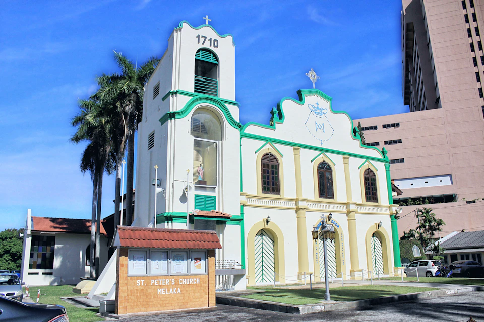 St. Peter's Church Melaka with green trim, white and yellow facade, tall bell tower, palm trees, and a sign reading "St. Peter's Church Melaka."