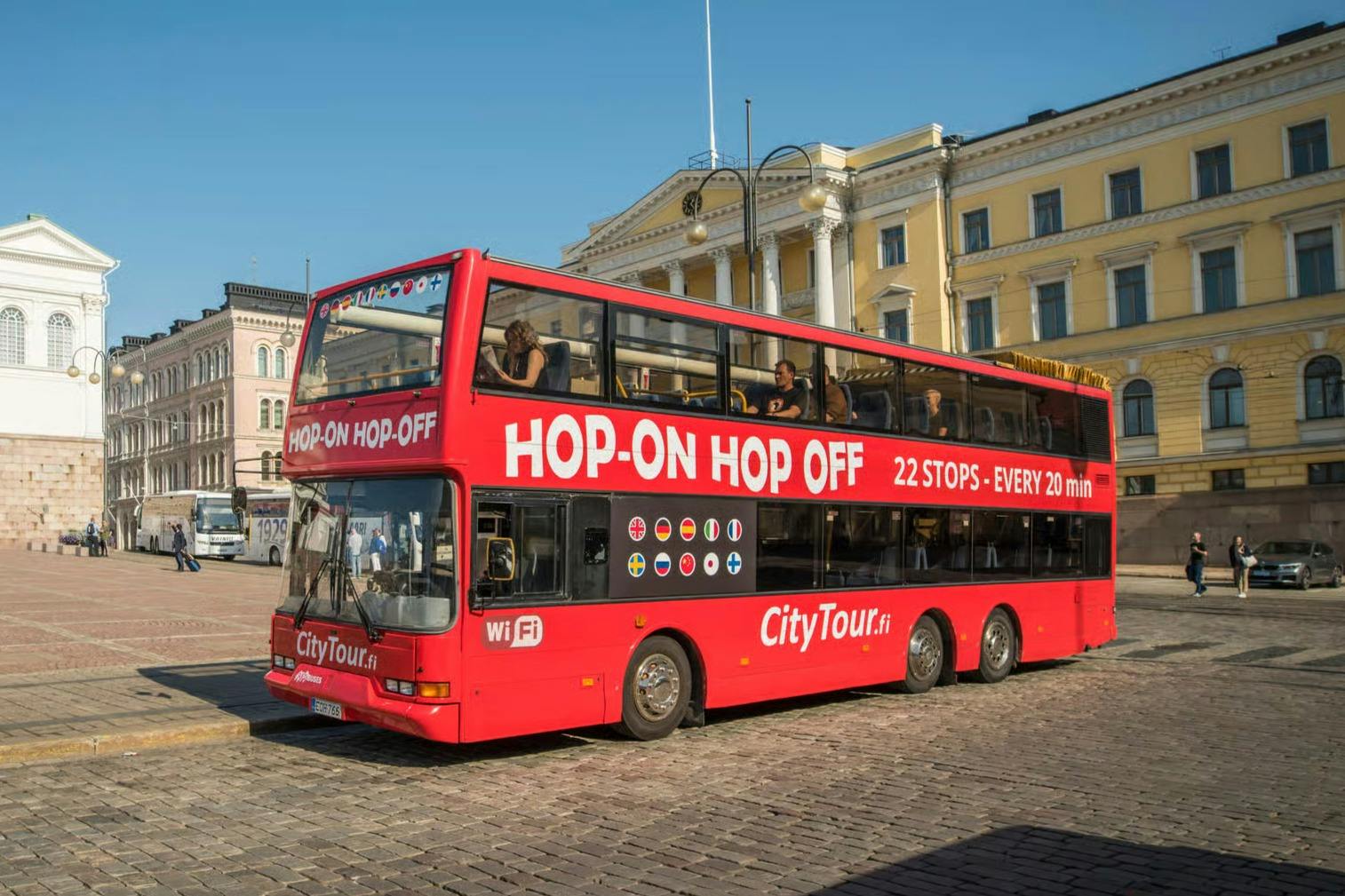 Un autobús vermell de dos pisos amb l'etiqueta "Hop-On Hop-Off" va aparcar en un carrer empedrat amb un edifici històric al fons.