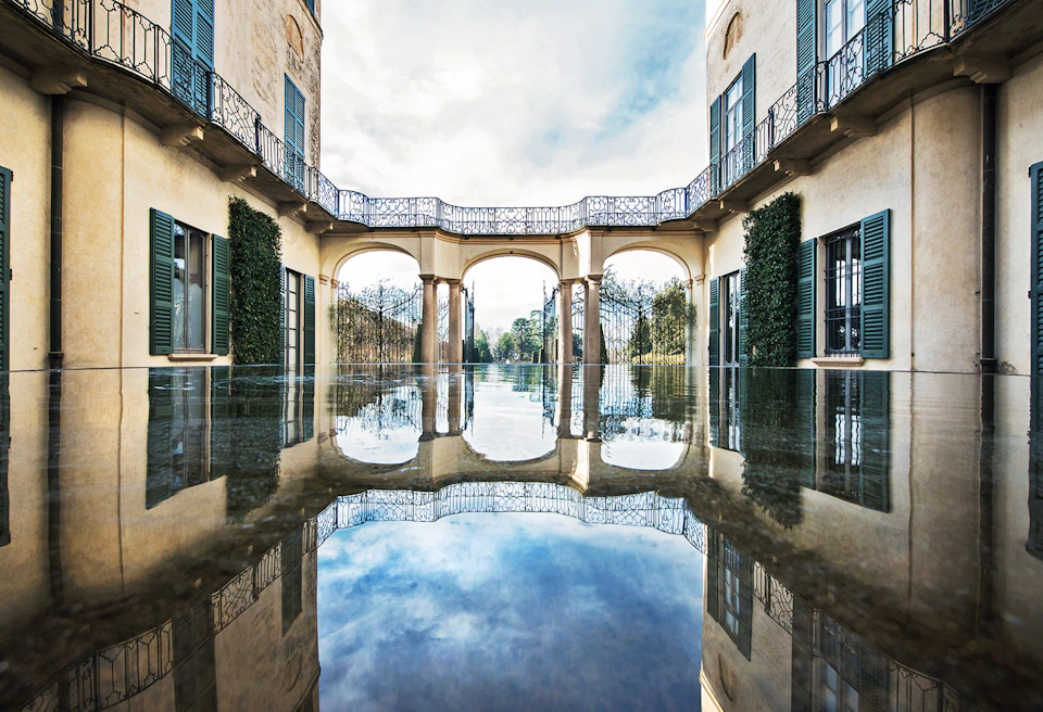 A courtyard with arched walkways, balconies, and a reflective water surface underneath a partly cloudy sky.
