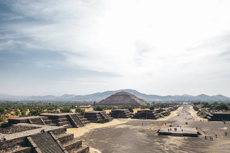 Ancient pyramids and structures on a wide avenue, with a mountainous landscape in the background under a partly cloudy sky.