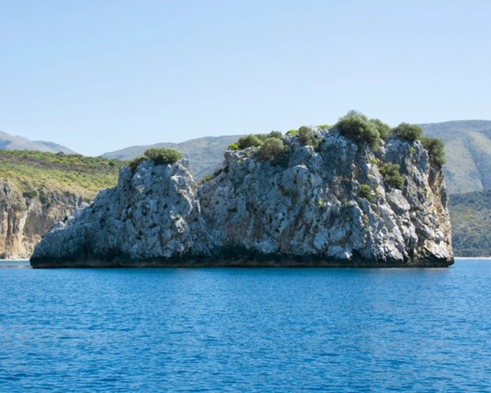 Rocky cliffs with sparse greenery rise from a calm blue sea under a clear sky. Additional hilly terrain is in the background.