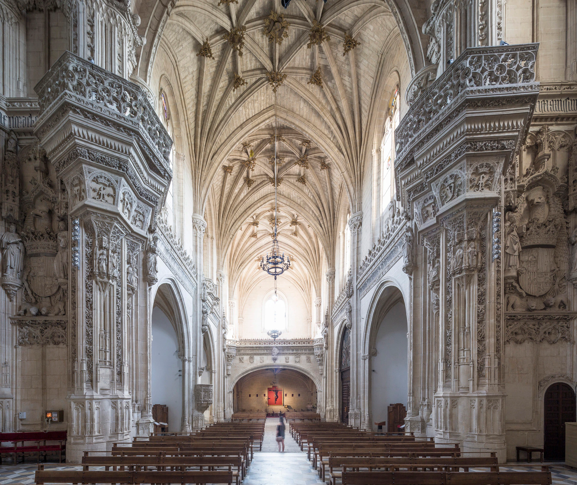 An ornate cathedral interior with stone carvings, vaulted ceilings, wooden pews, and a person walking down the central aisle.