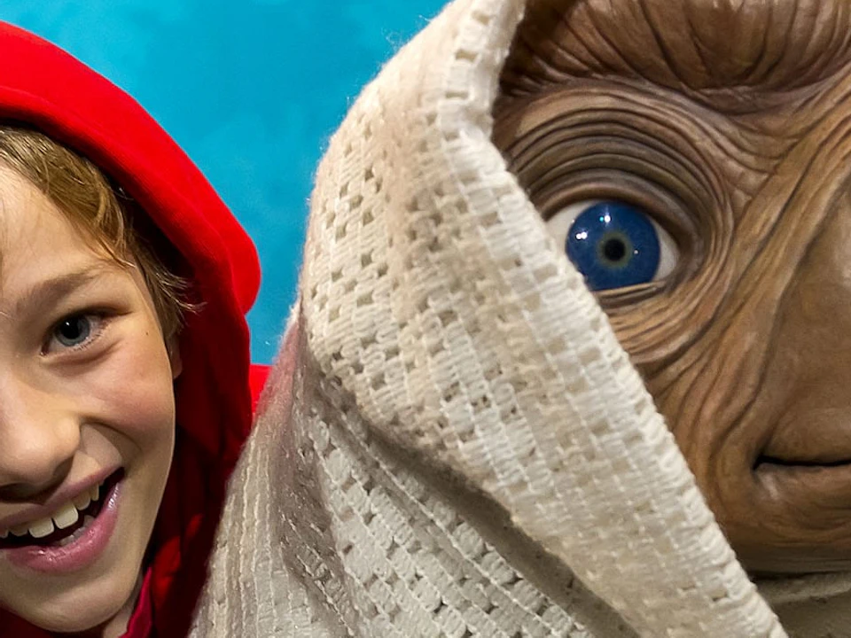 Smiling child in a red hoodie next to an alien figure wrapped in a white blanket, set against a blue background with silhouettes of trees.