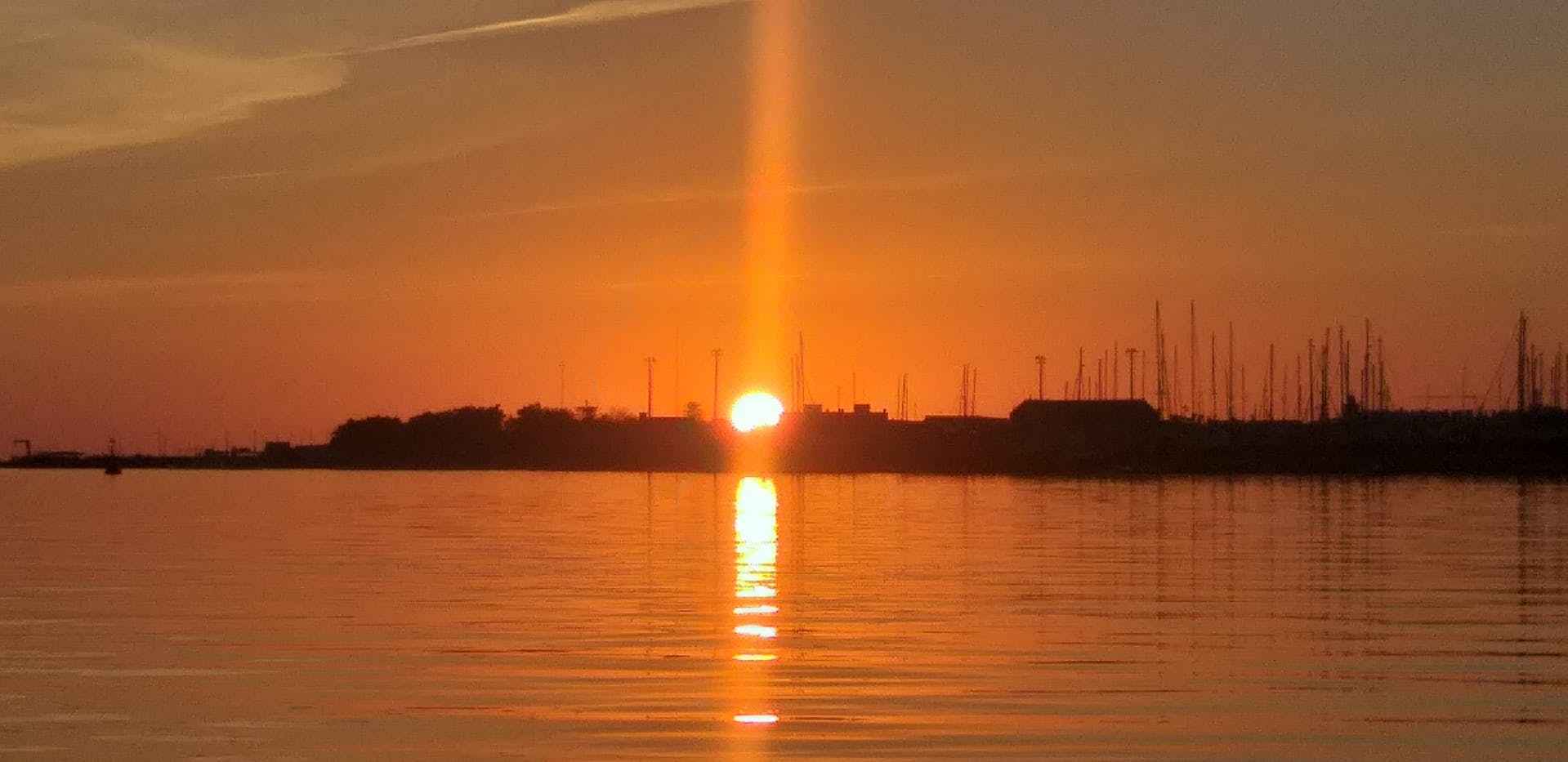Закат над спокойной водой с солнцем частично за горизонтом силуэтных зданий и мачт, создавая вертикальное отражение света.