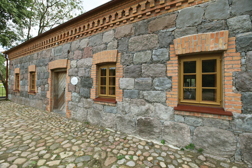 A stone building with a wooden door and two windows framed by brick, situated on a cobblestone pathway.