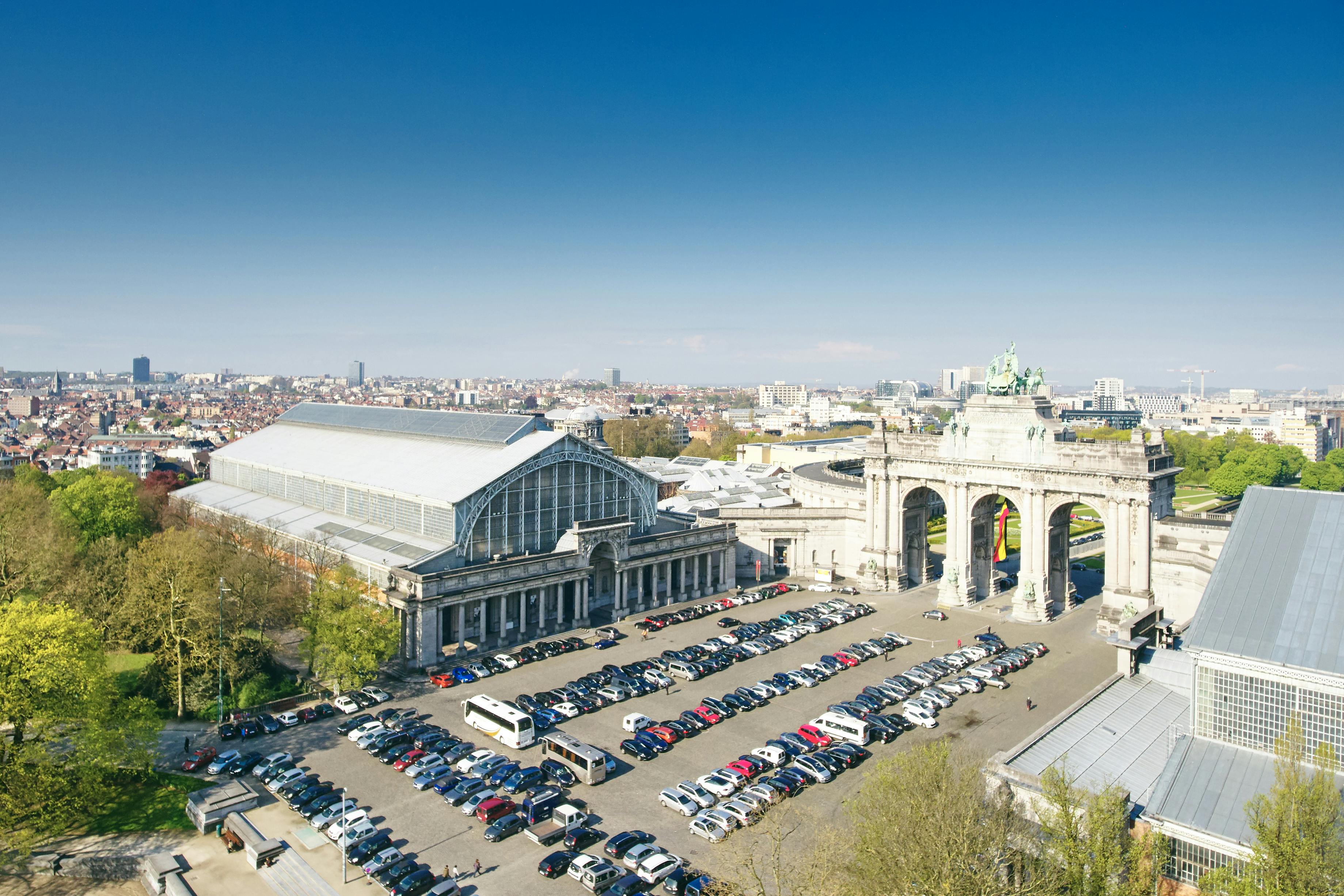 Vue grand angle d'un grand bâtiment à toit de verre à côté d'un monument en forme d'arche, avec un parking bondé et un paysage urbain à l'arrière-plan.