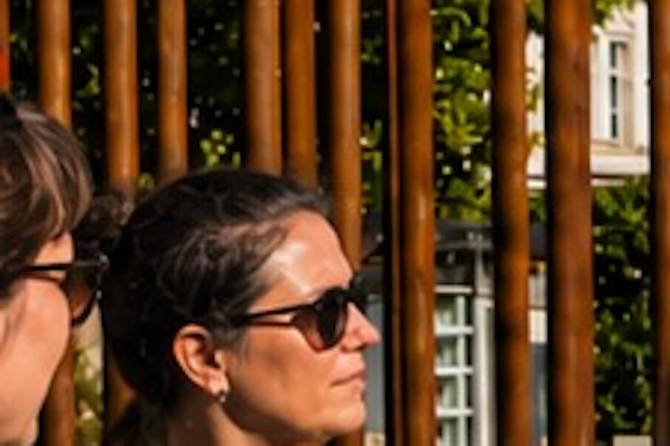 A group of people chatting near a tall, rusty metal fence in a green park on a sunny day with scattered clouds.