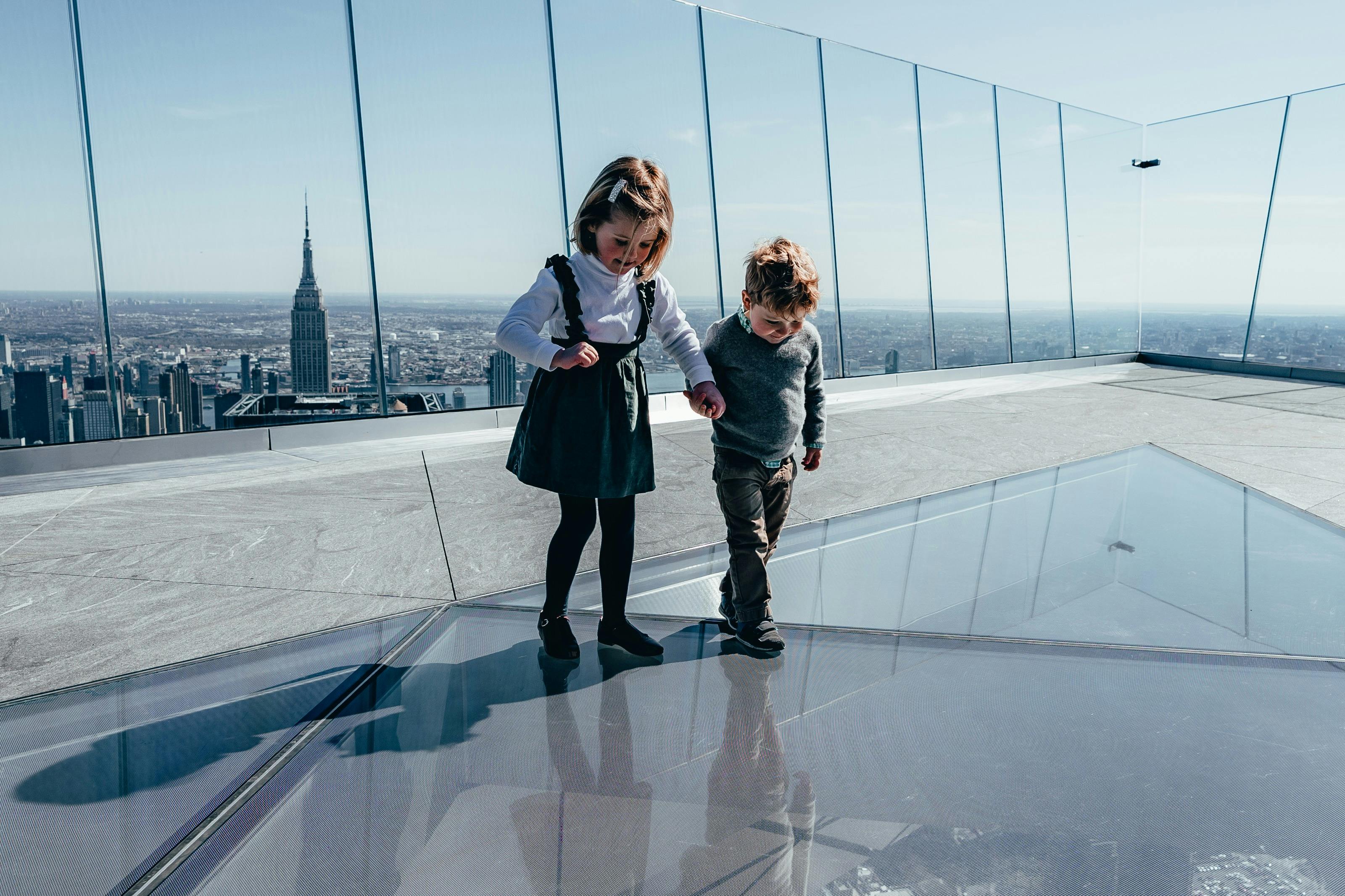 Dvě děti jdou ruku v ruce po skleněné podlaze na vrcholu mrakodrapu s panoramatem včetně Empire State Building za nimi.