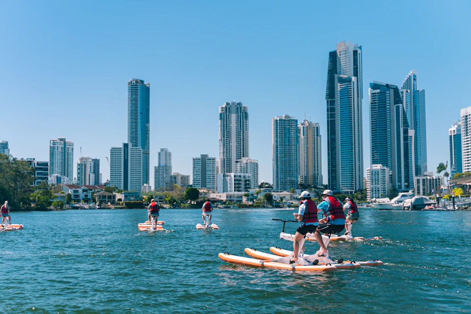 People paddleboarding on a waterway with a city skyline of tall buildings in the background on a sunny day.
