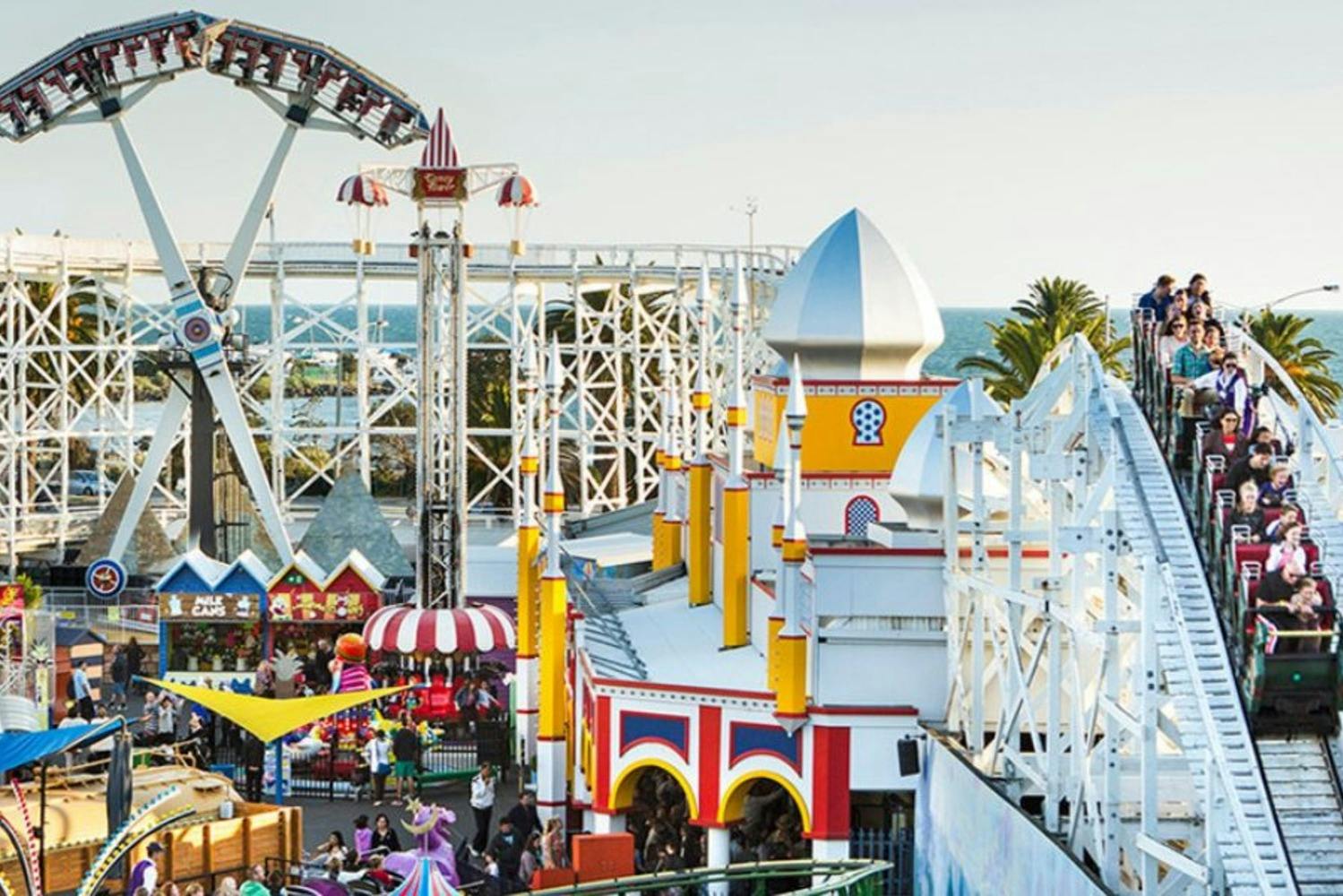En levende forlystelsespark med rutsjebaner, forlystelser, farverige bygninger og folk, der nyder seværdighederne. Havet er synligt i baggrunden.