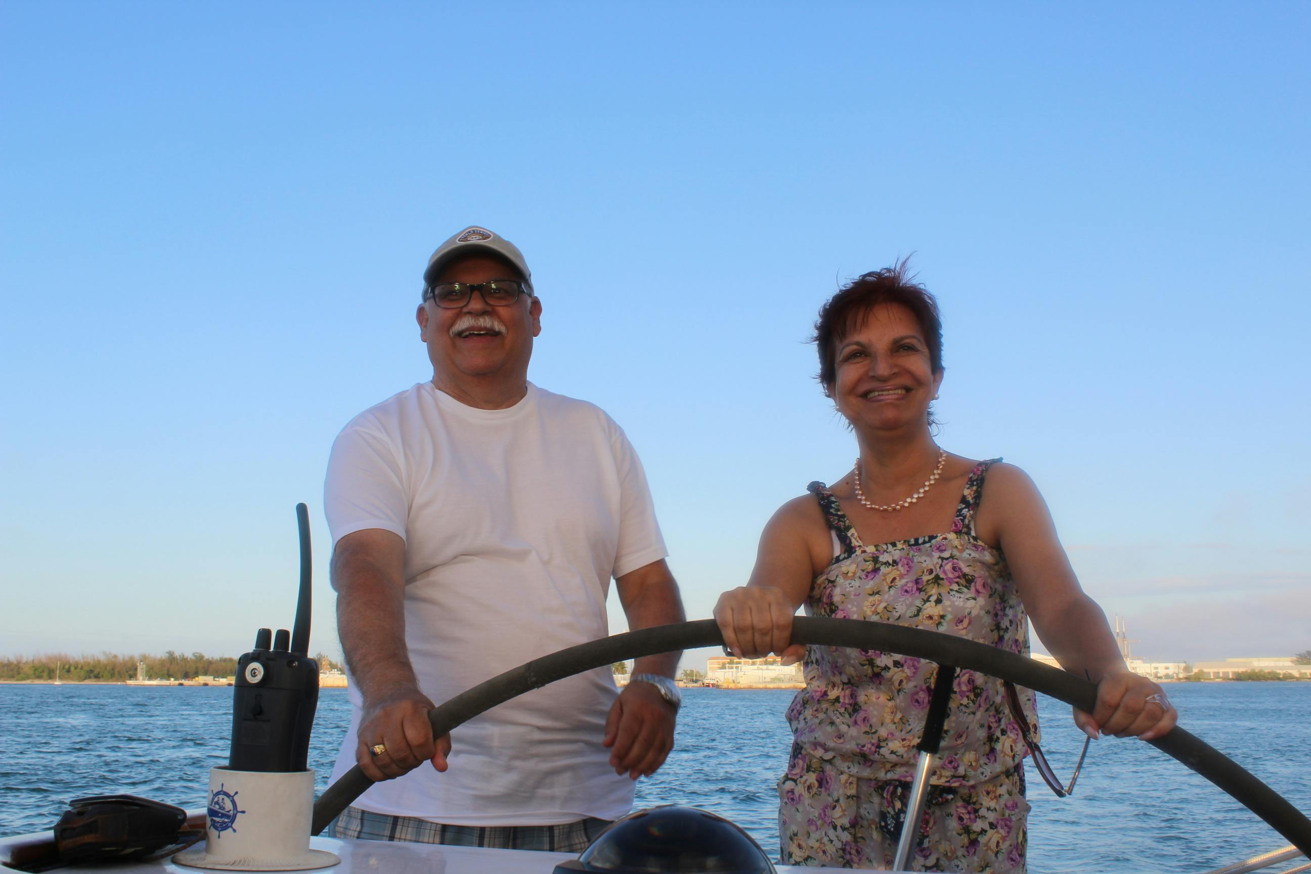 Un homme et une femme, tous deux souriants, dirigent un bateau sur une eau calme, sous un ciel bleu clair.
