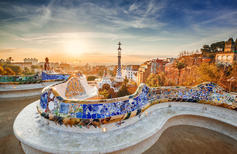 View of mosaic-tiled serpentine bench, buildings, and cross-topped tower in Park Güell, Barcelona, with a sunset sky.