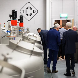 Människor i kostymer inspekterar stora, cylindriska metalltankar med maskiner. Väggen har "TCD" -logotypen i svart.