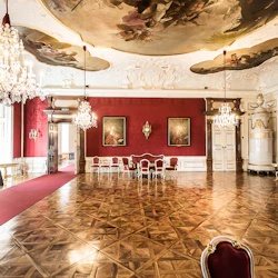 Genomarbetat rum med röda väggar, ljuskronor, utsmyckade takmålningar, polerat trägolv, röd matta och vintage-möbler.