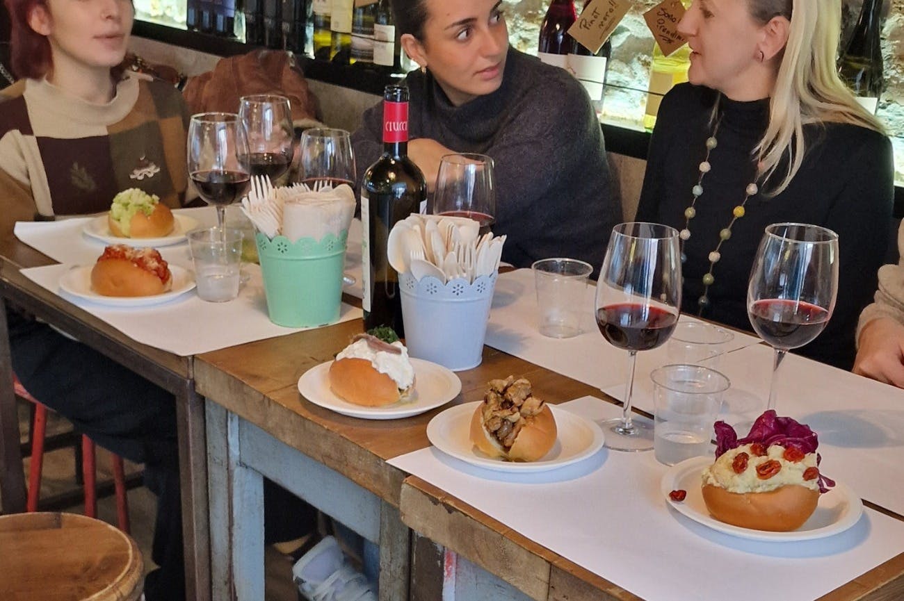 Two women conversing at a table with wine glasses, bottled wine, plates of food, and napkin holders in a rustic setting.