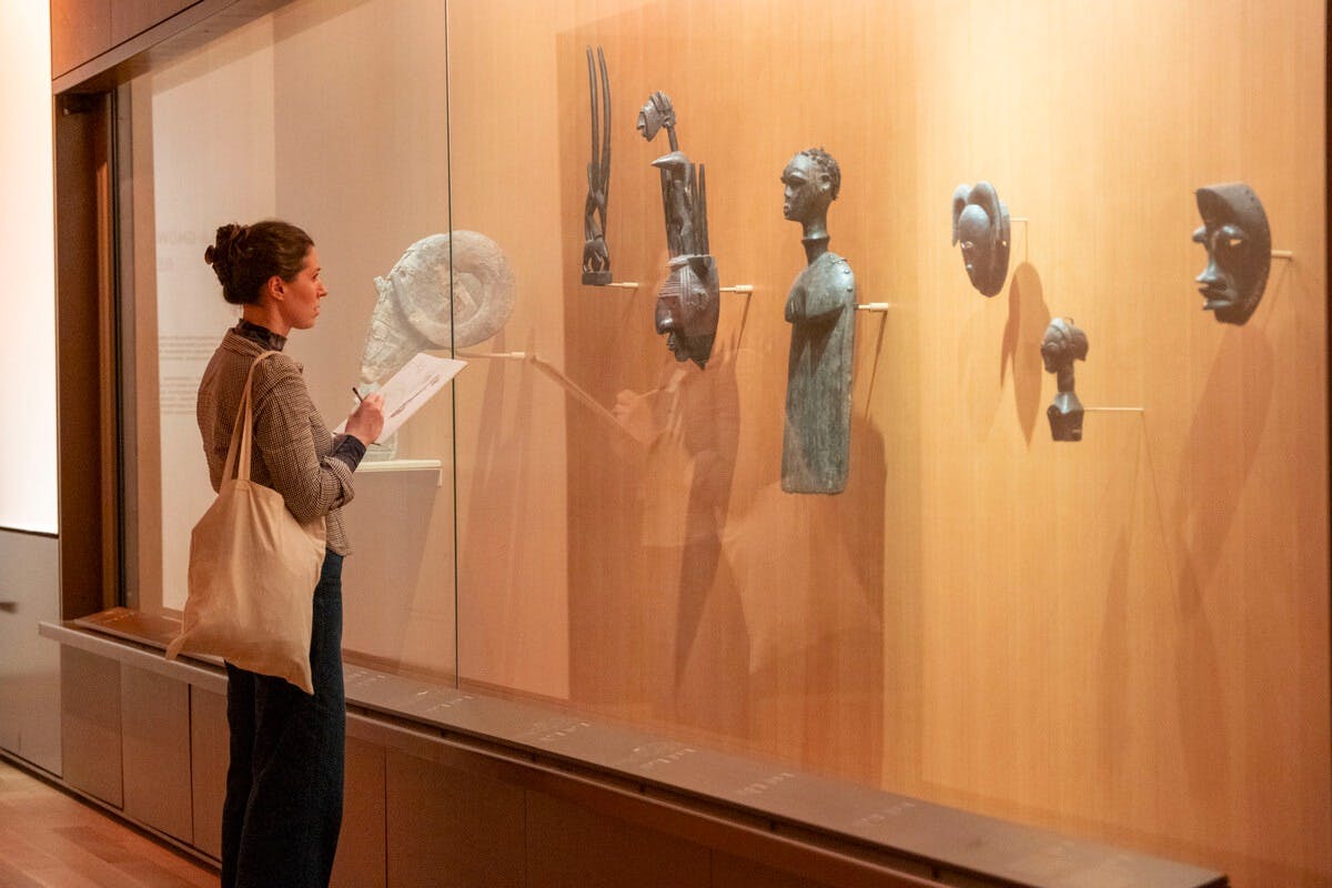 Eine Frau betrachtet die in einem Museum ausgestellten Skulpturen, hält eine Broschüre in der Hand und trägt eine Umhängetasche.