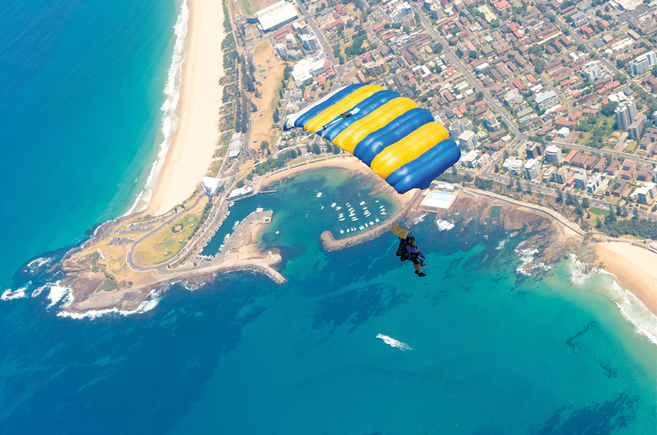 Skydiver descending with a yellow and blue parachute over a coastal city, showing beach, marina, and urban landscape below.