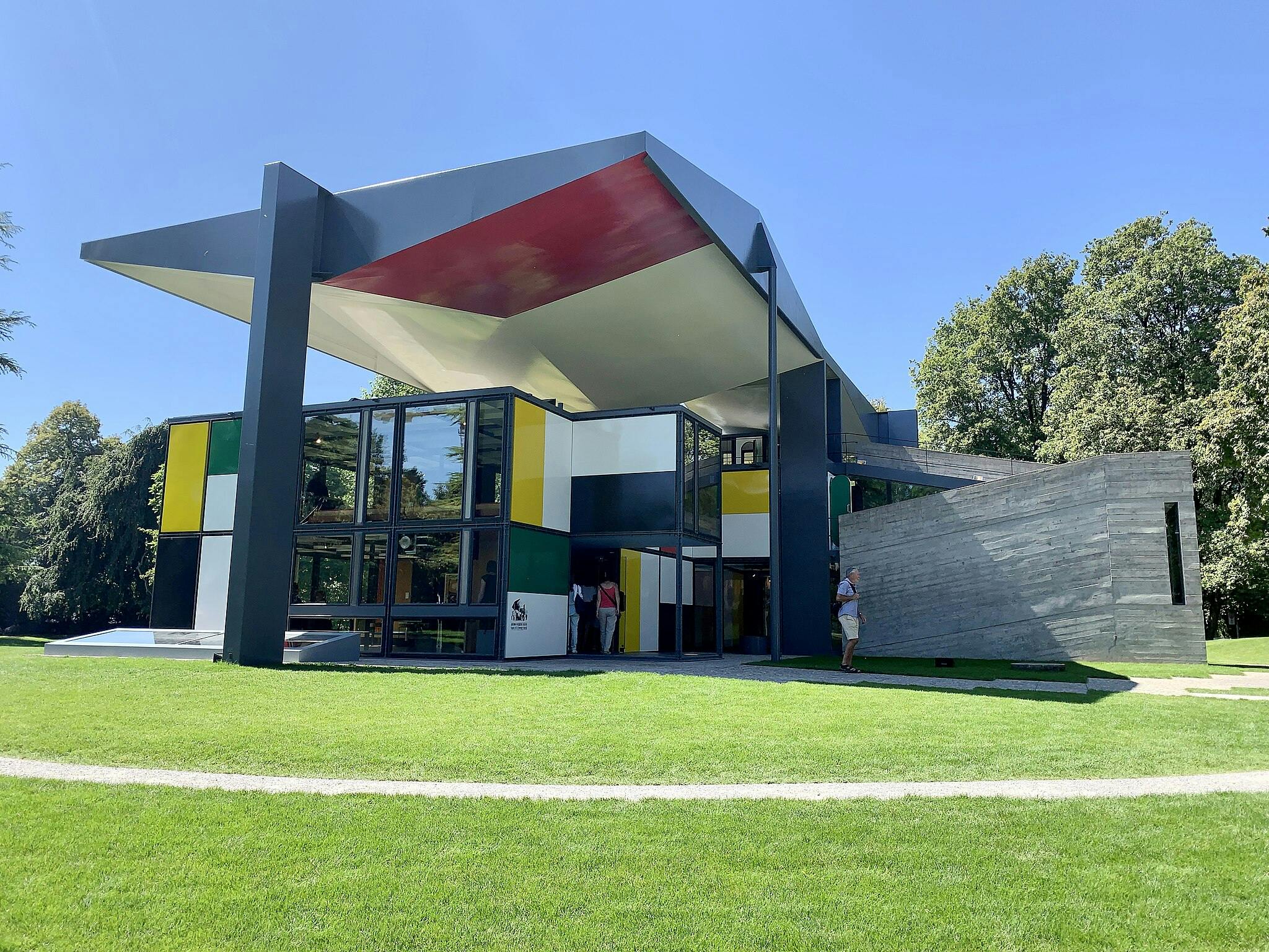 カラフルなパネル、大きな窓、緑の芝生に張り出した屋根を備えた、幾何学模様のモダンな建物。