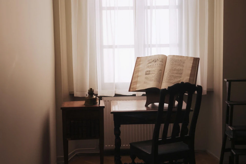 A desk and chair by a window with sheer curtains, featuring an open book on a stand and a small side table nearby.