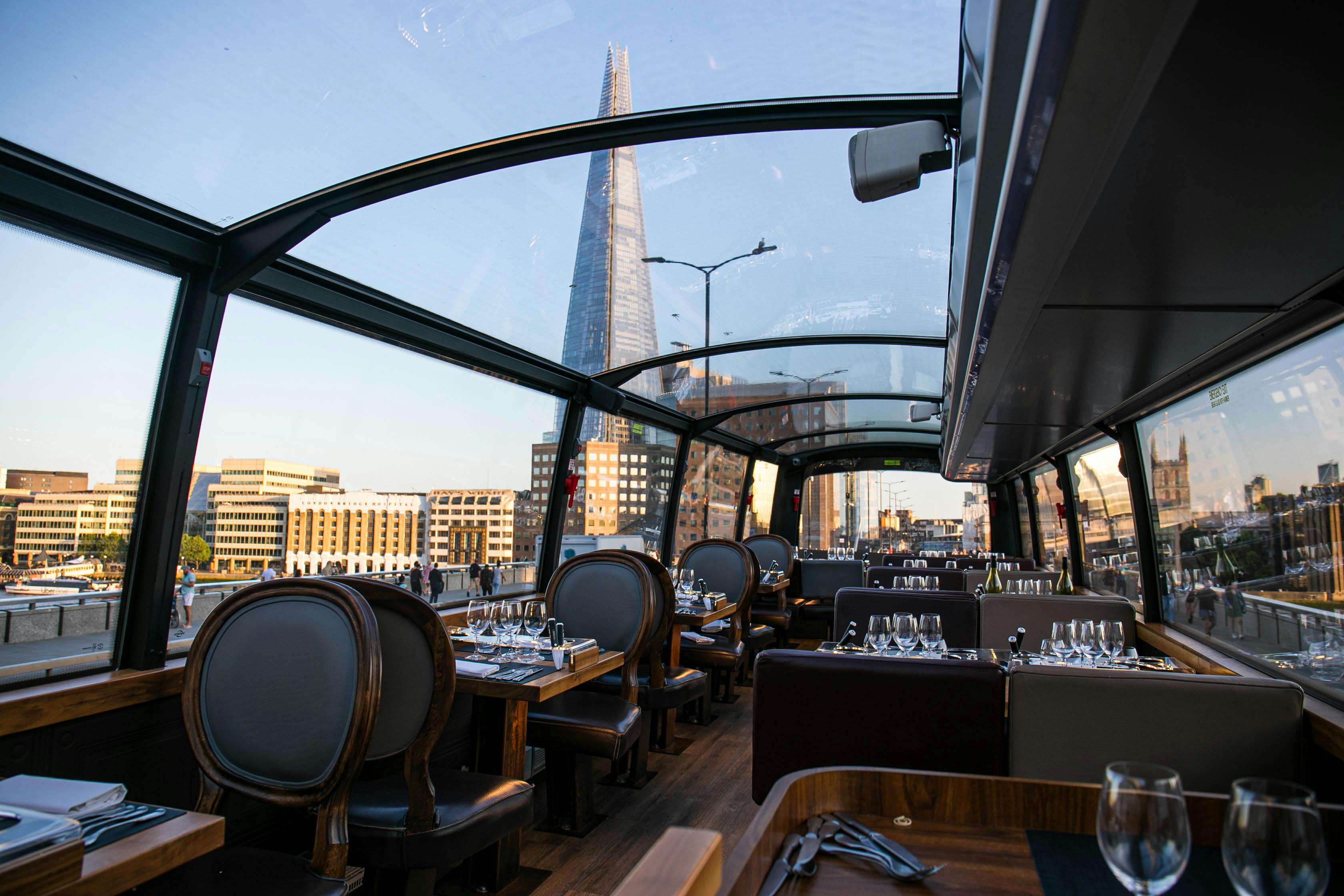 Interior of a glass-roofed dining area with set tables and chairs, overlooking city buildings and the Shard skyscraper in the distance.