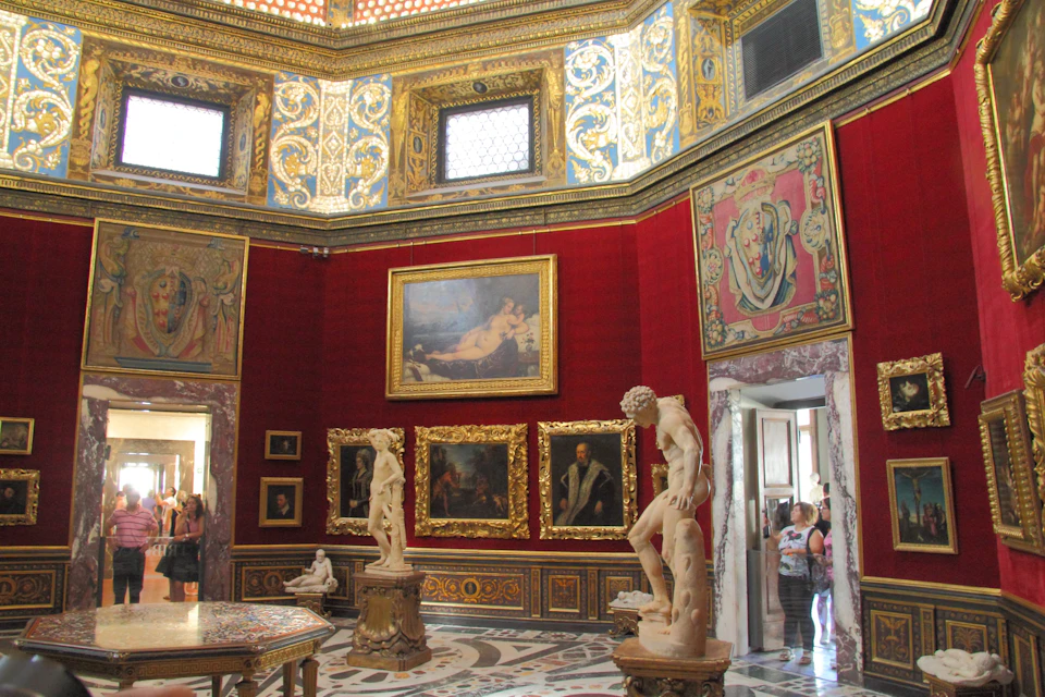 An ornate museum gallery with red walls, gold-framed paintings, and classical sculptures on pedestals. Visitors observe the art.