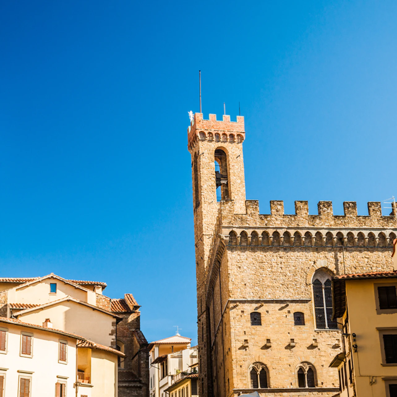 Historic stone building with a crenelated tower against a clear blue sky; surrounding buildings with tiled roofs.