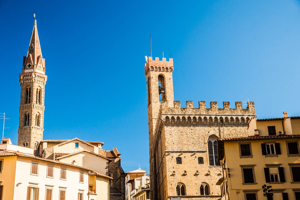 Historic stone building with a crenelated tower against a clear blue sky; surrounding buildings with tiled roofs.