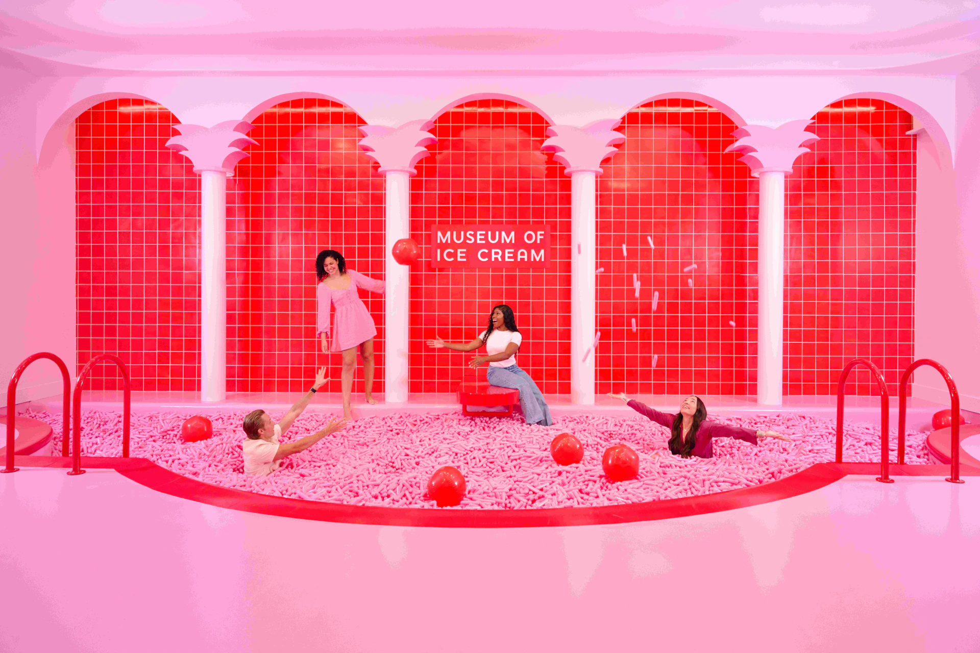 Four people enjoy posing and playing with red balls in a pink ball pit under an archway labeled "Museum of Ice Cream."