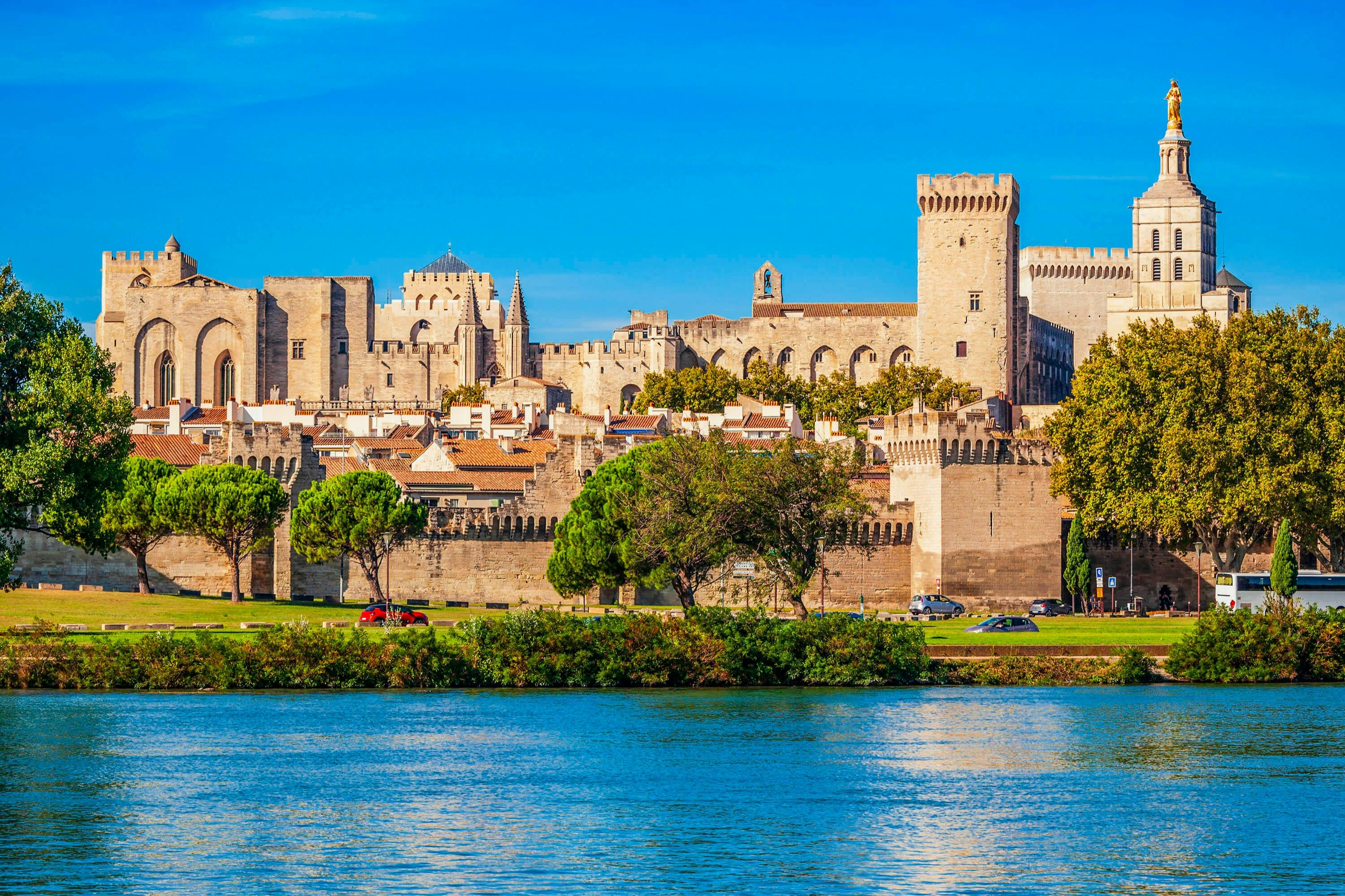 A historic fortress with towers and walls viewed from across a river, surrounded by trees and houses under a clear blue sky.