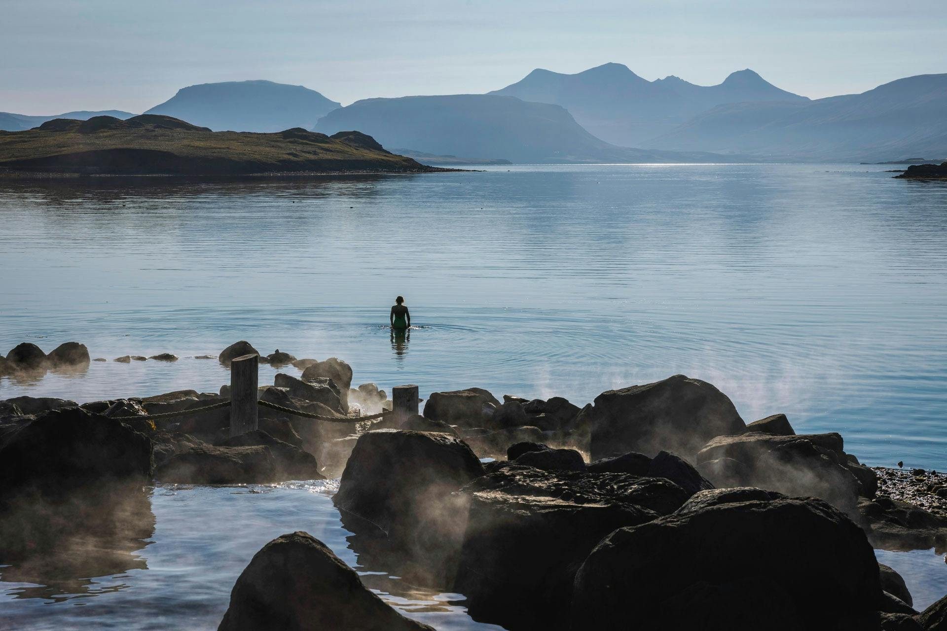 一个孤独的人站在一个湖泊的平静水面上，周围环绕着岩石，浓雾升起，背景是山脉。