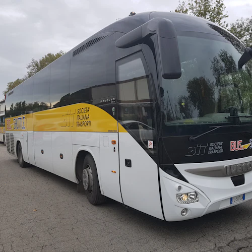 En vit och gul buss märkt "Societa Italiana Trasporti" och "Bus Shuttle" parkerade utomhus med en annan buss i bakgrunden.