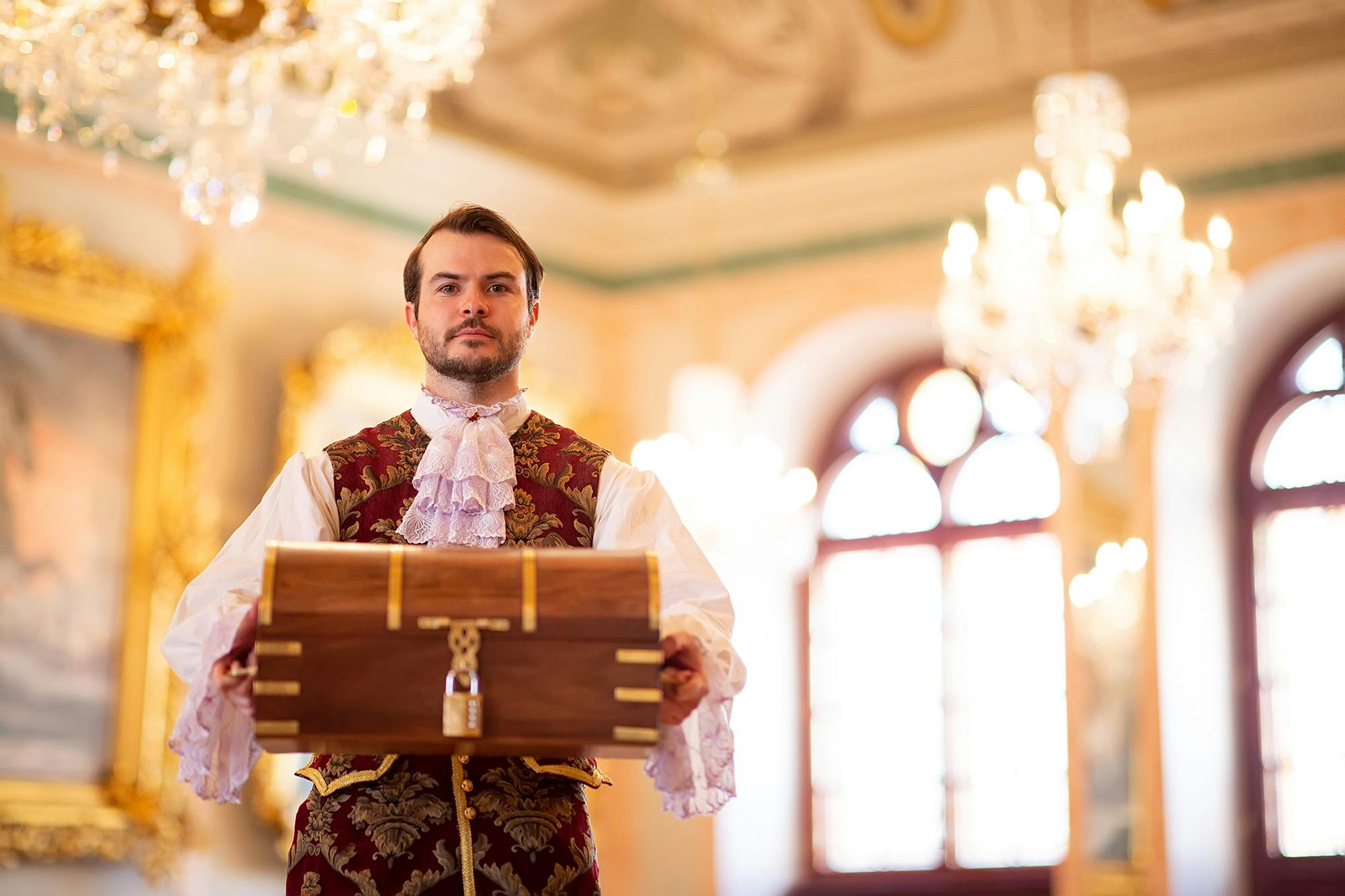 穿着古装的男子在华丽的房间里拿着一个木箱，里面有吊灯和框架艺术品。