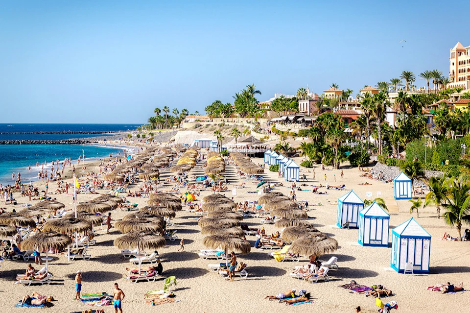 Crowded beach with straw sun umbrellas, sunbathers on lounge chairs, and cabanas; palm trees and buildings in the background.