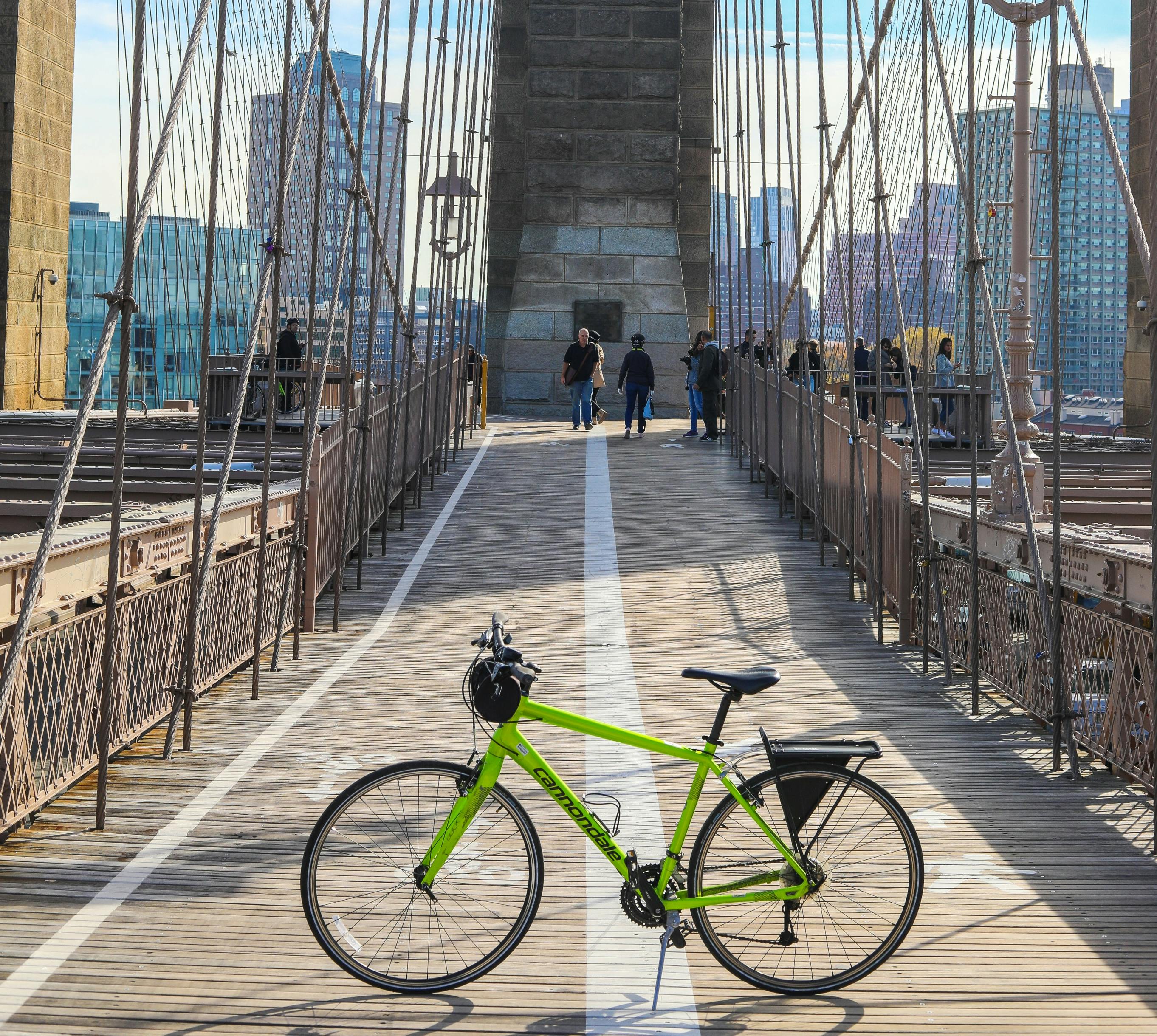 Una bicicletta verde brillante è parcheggiata sulla passerella di legno del ponte di Brooklyn con persone che camminano sullo sfondo ed edifici della città visibili.