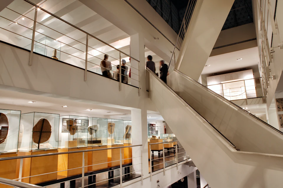 Modern museum interior with glass display cases and artifacts. People are viewing exhibits on the upper level near a staircase.