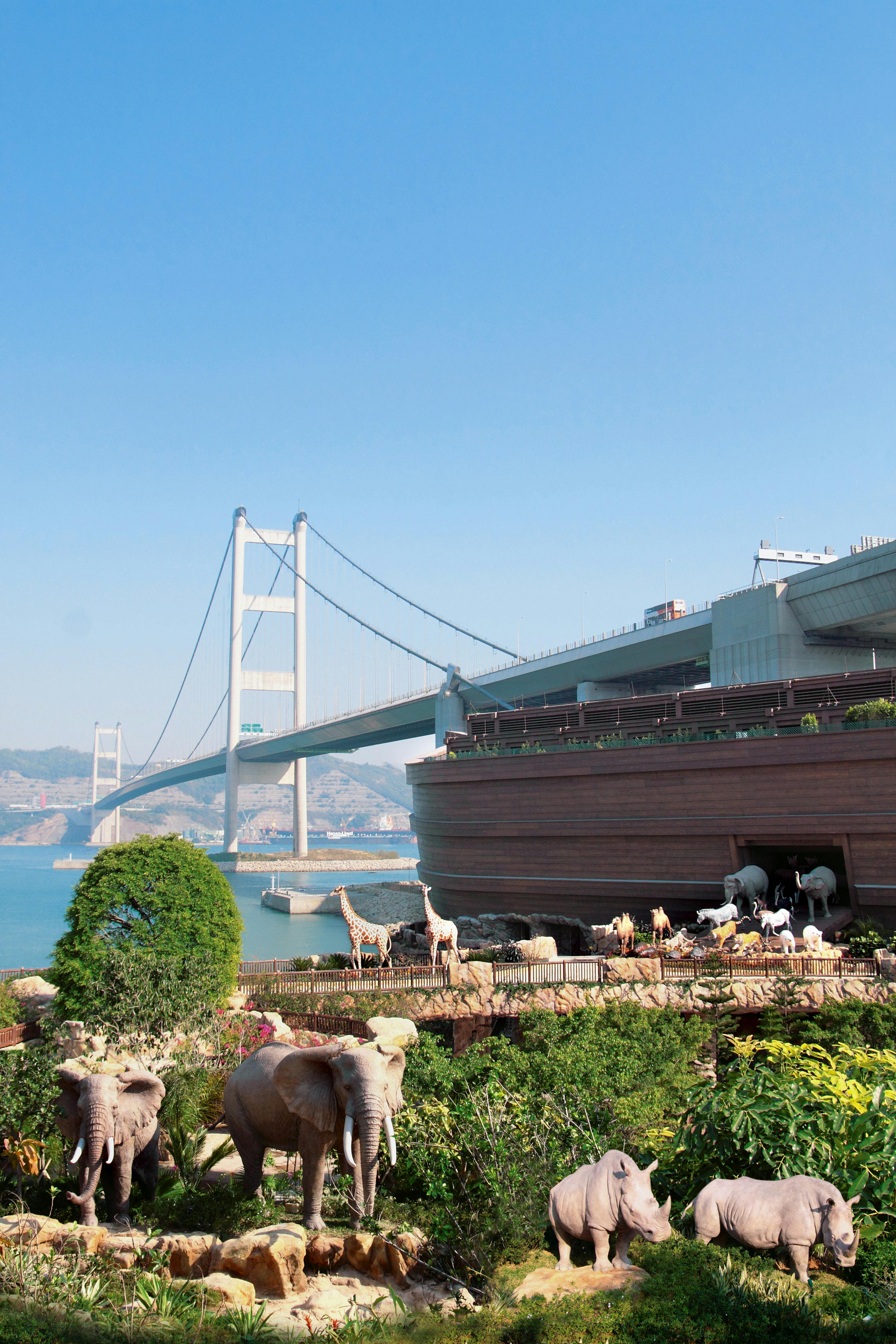 Большое деревянное здание, напоминающее Ноев ковчег, окруженное различными скульптурами животных, рядом с подвесным мостом над водой.